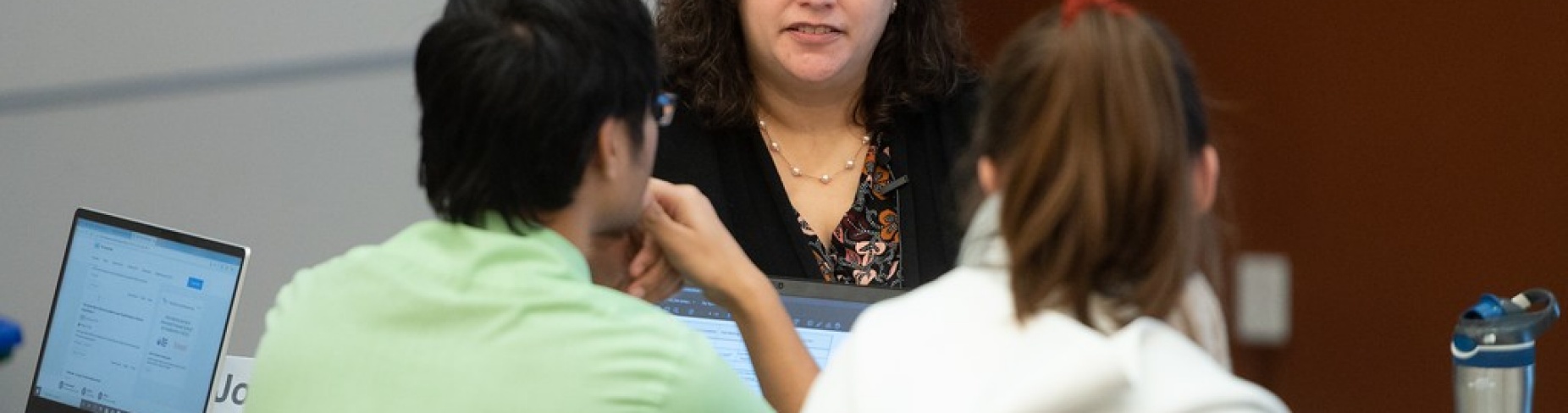 Sabrina Casucci, associate professor of teaching, sits in front of two students who have their backs turned to the camera at a table in a classroom. 