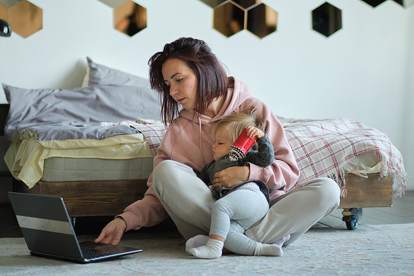 A young mother sits on the floor holding her squirming baby as she tries to do work on a laptop computer. 