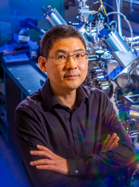 Researcher Hao Zeng in the lab next to a long machine with many metal chambers and working parts. The photo is shot artistically, with blue and rainbow light effects. 