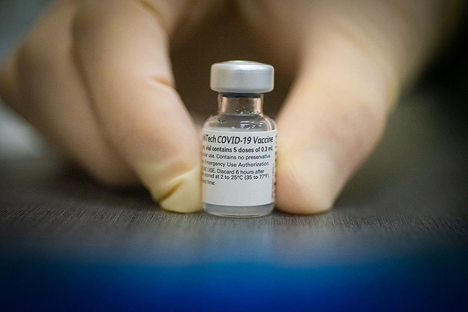 Close up of gloved fingers holding a vial of Pfizer BioNTech Covid 19 vaccine. 