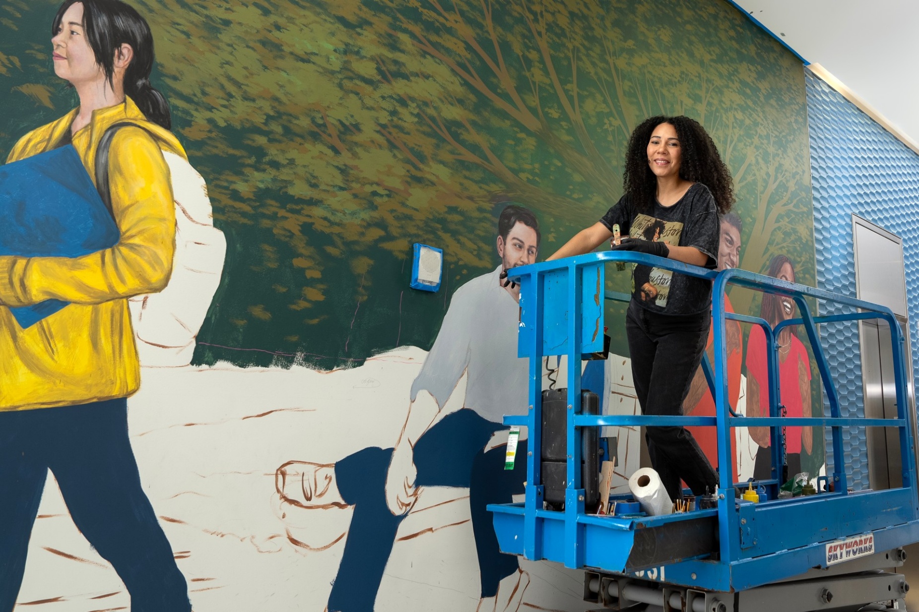 Julia Bottoms stands on scissor lift in front of half finished mural. 