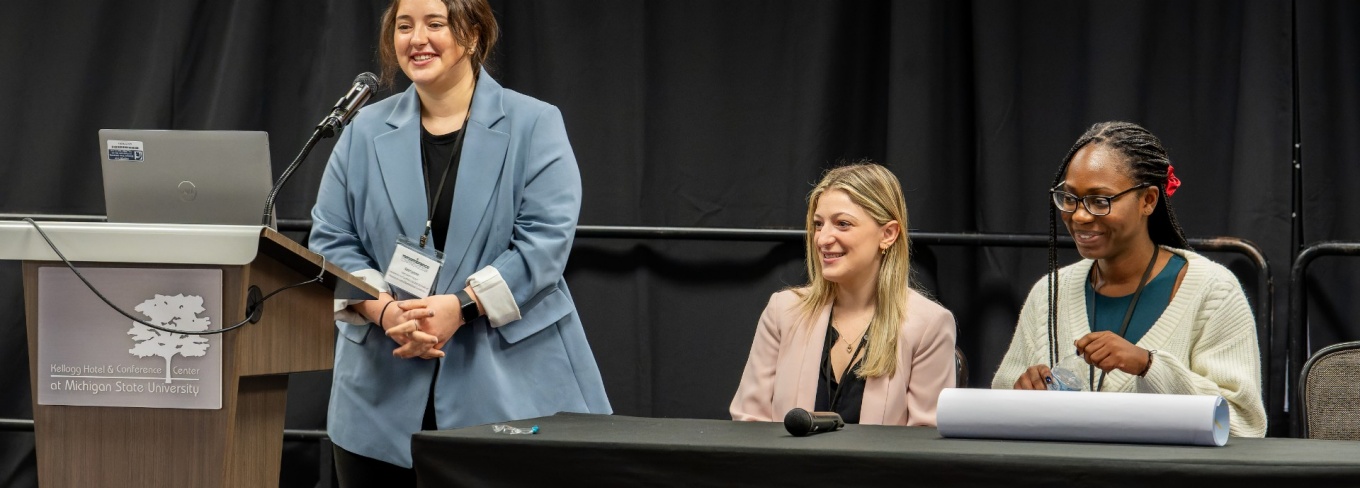 L to R: Kerryann Koper standing at podium, Miranda Berkebile and Koulako Kaba are seated. 