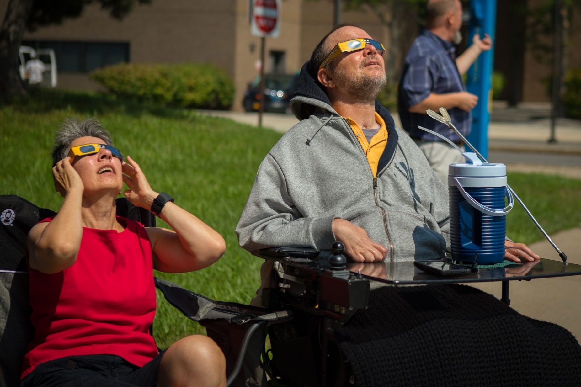 Two people seated on a lawn, one is in a wheelchair, looking up and wearing eclipse glasses. 