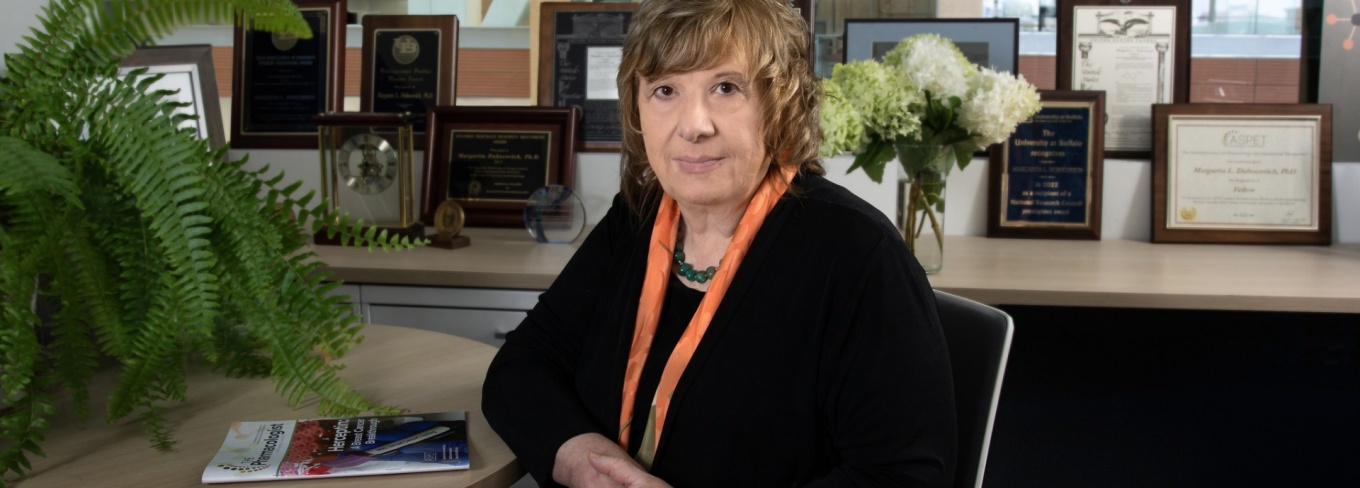 Margarita Dubocovich is sitting at a desk, surrounded by awards and a plant. 