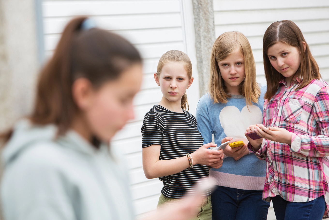 An adolescent girl in the foreground looking at a phone while another group of adolescent girls, also using phones, look at her in an unfriendly way. 