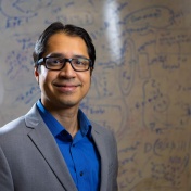 Souma Chowhury stands in front of a white board covered with words and equations. 