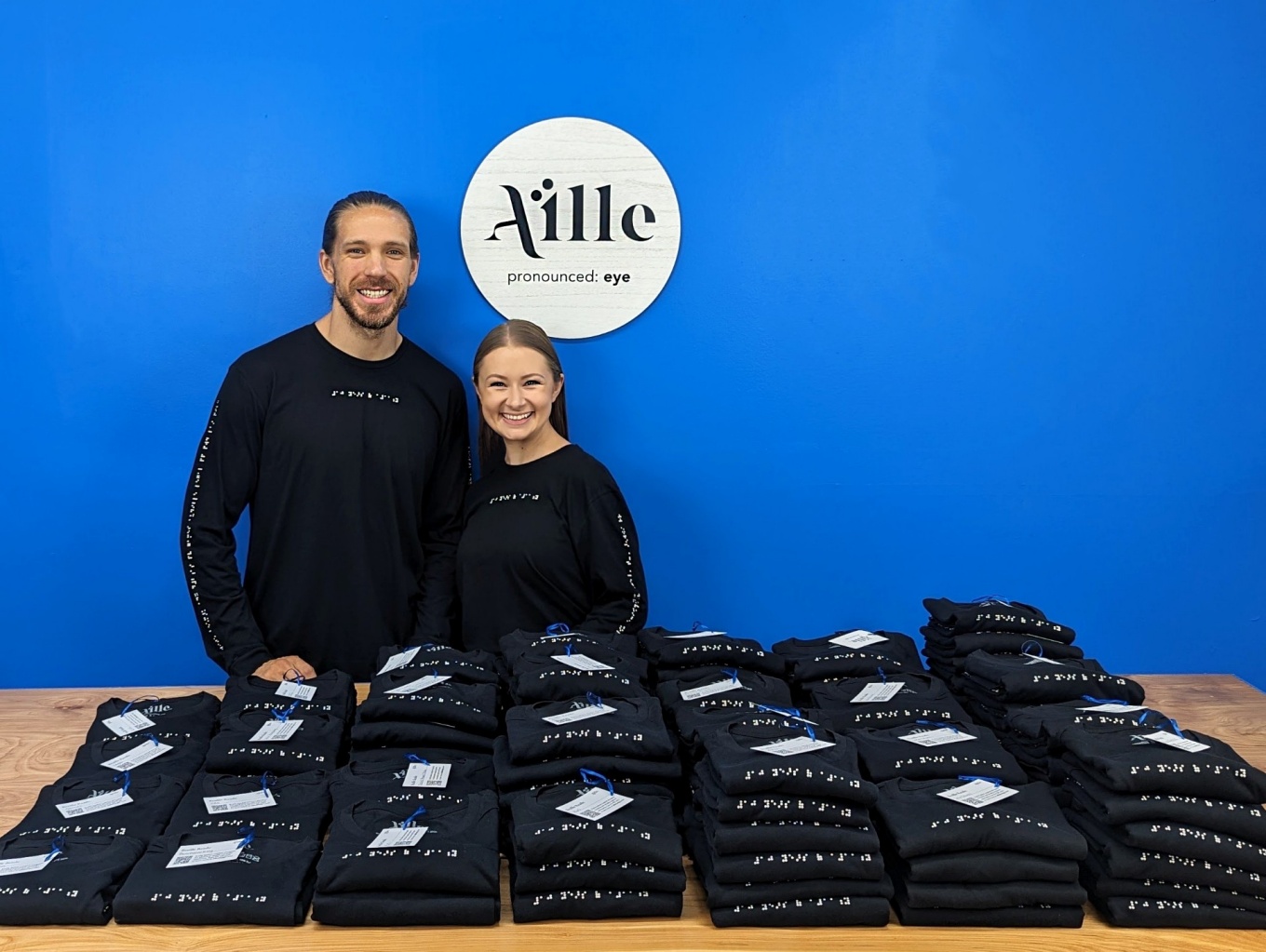 From left, Jake Walsh and Alexa Jovanovic, Aille Design team, standing behind a table of folded t-shirts bearing their braille designs. 
