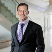 Photo of Elad Levy in suit and tie standing in medical school atrium with staircase behind him. 