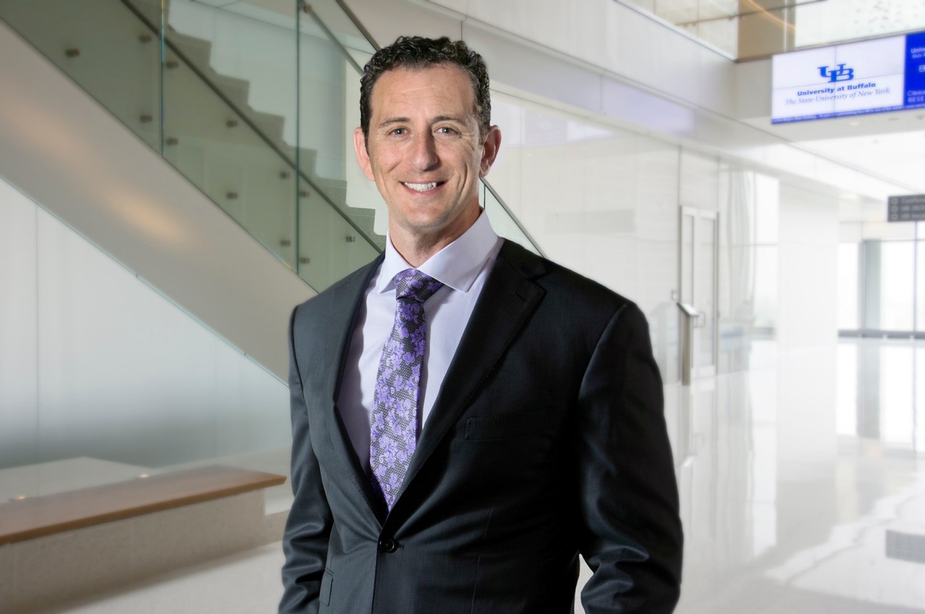Photo of Elad Levy in suit and tie standing in medical school atrium with staircase behind him.