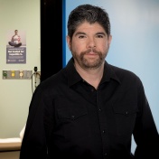 Anthony Martinez is standing in a hallway outside an exam room where there is a poster on the wall about pregnancy and hepatitis C. 
