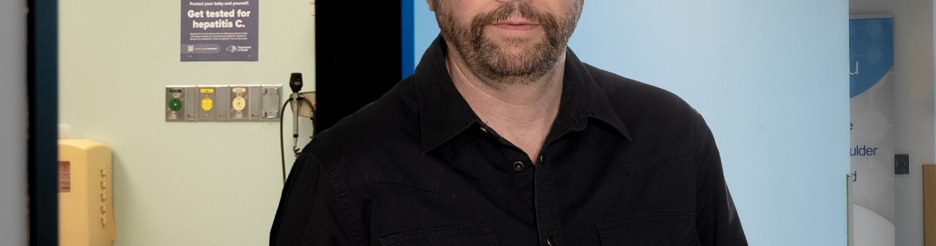 Anthony Martinez is standing in a hallway outside an exam room where there is a poster on the wall about pregnancy and hepatitis C.