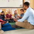 A teacher and students sit on the floor, each with a tablet. 