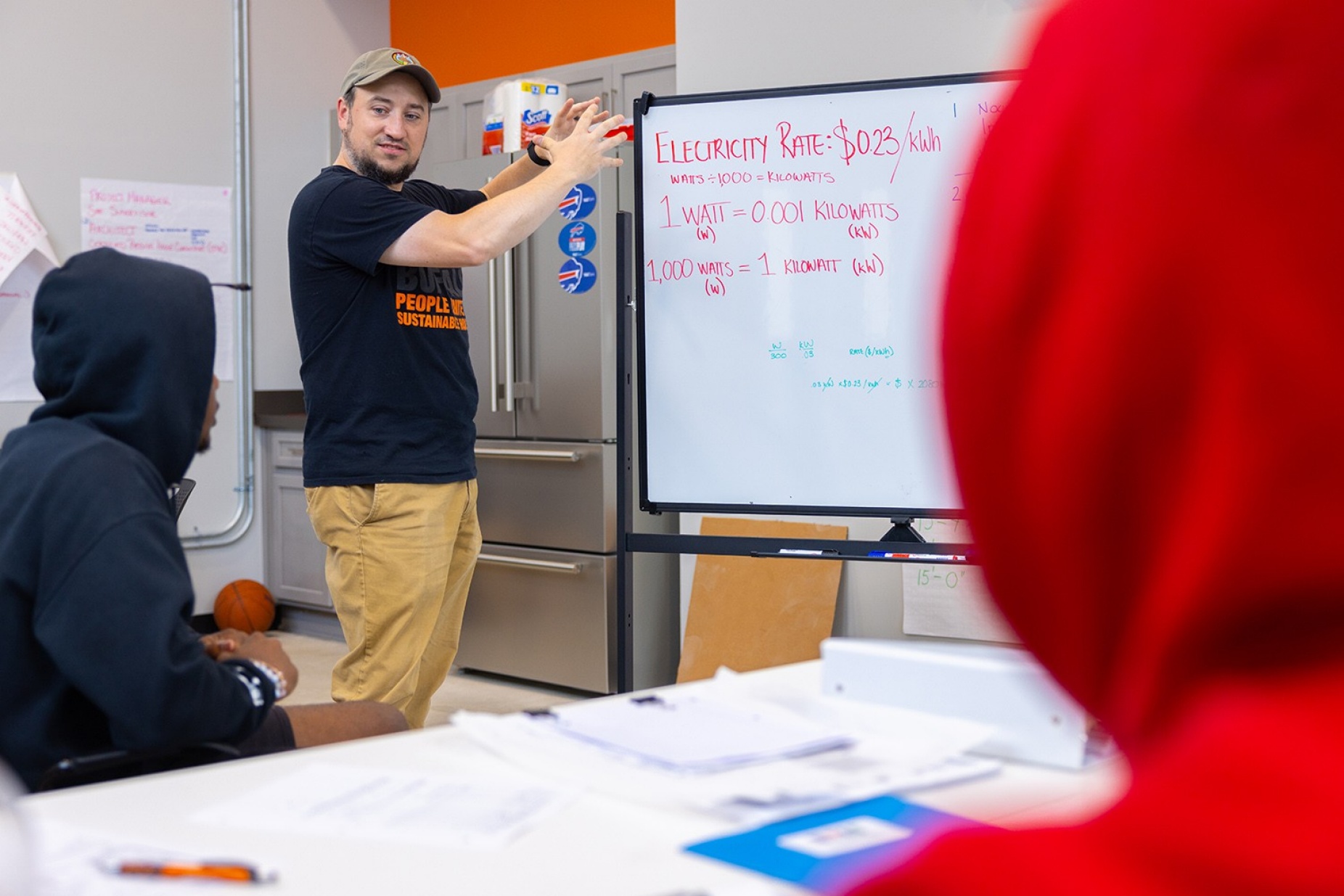 Male wearing a baseball cap standing in front of a whiteboard as he teaches two students. 