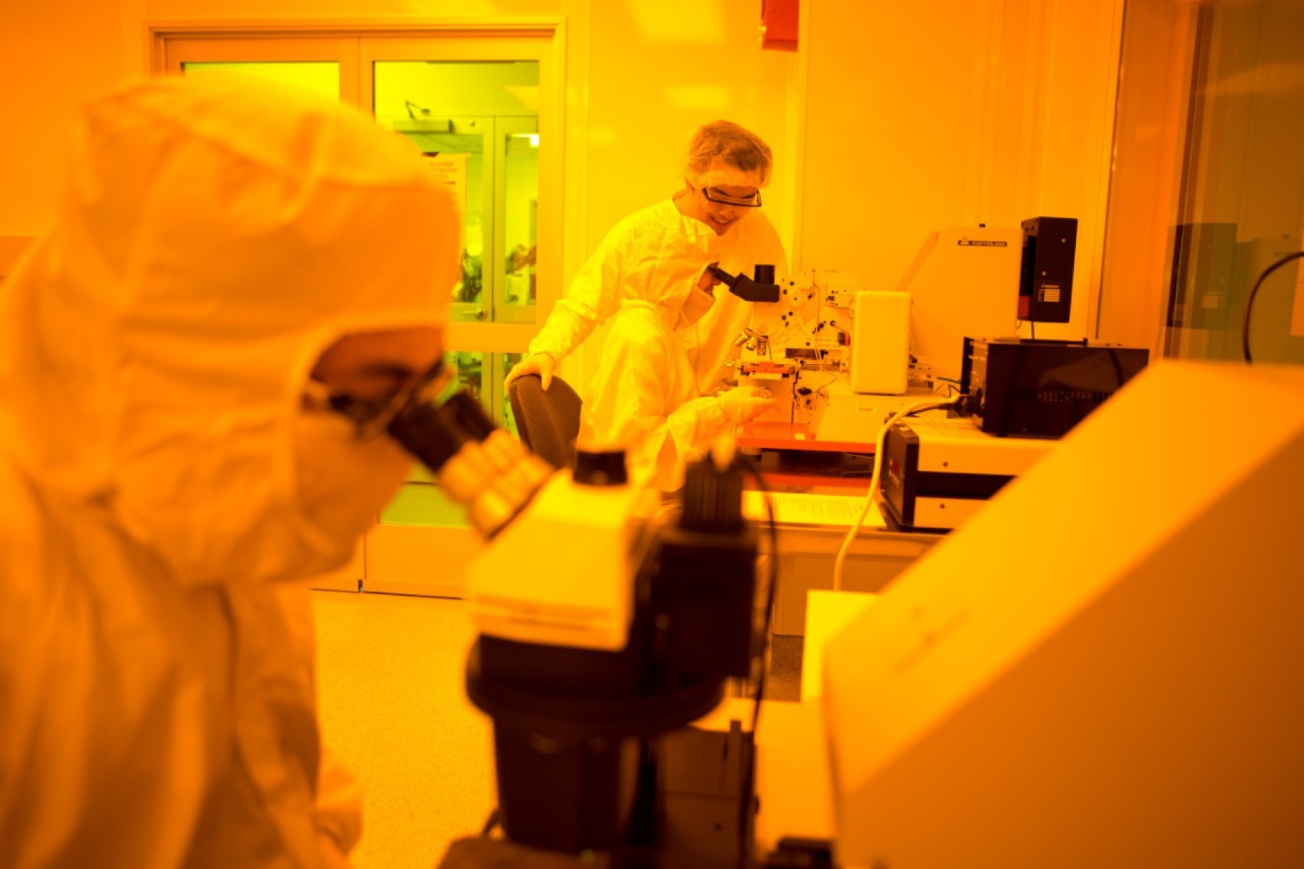 Inside the clean room at Davis Hall. Two students look through microscopes while one student looks on. They are wearing protective white lab suits. 