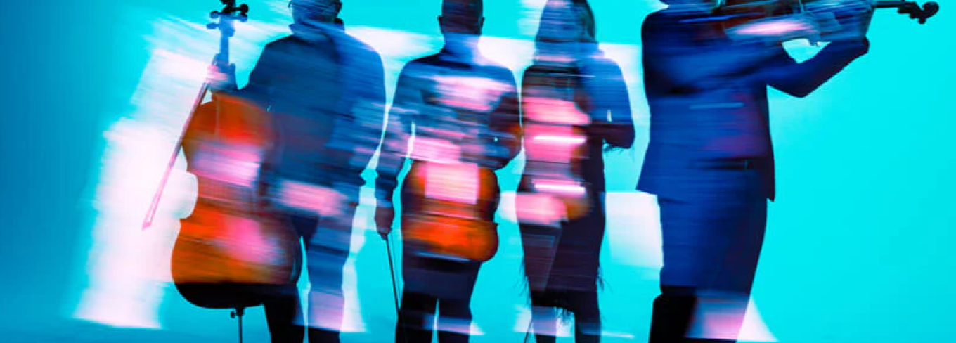 Blurred photo with a blue background of the four members in the Vitamin String Quartet posing with their string insturments. 