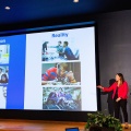 A person delivers a presentation standing next to a large screen on stage. 