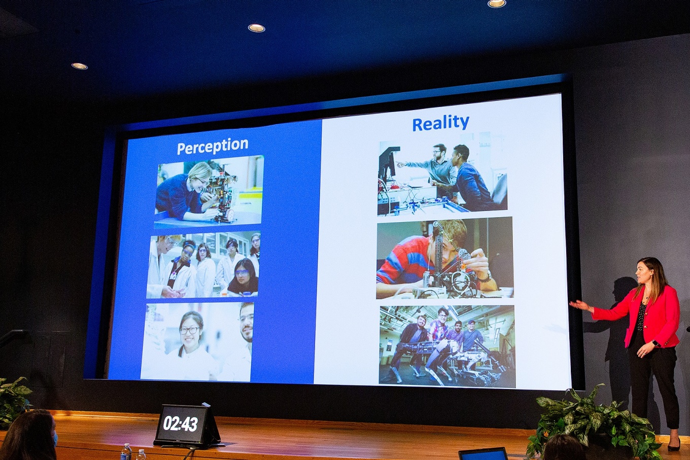 A person delivers a presentation standing next to a large screen on stage. 