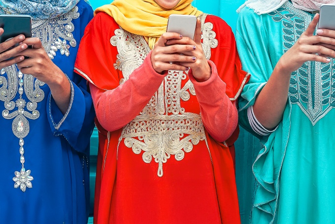 Three women wearing ornate South Asian-style dresses and looking at smart phones. 
