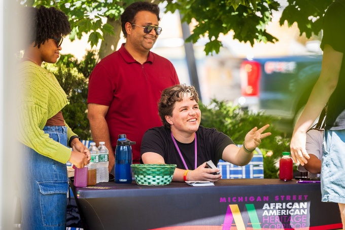 Social Impact fellow Cayden Edwards seated at an informational table display at the Mrs. Frances Nash's Garden Basket Farmer's Market in the Michigan Street African American Heritage Corridor.