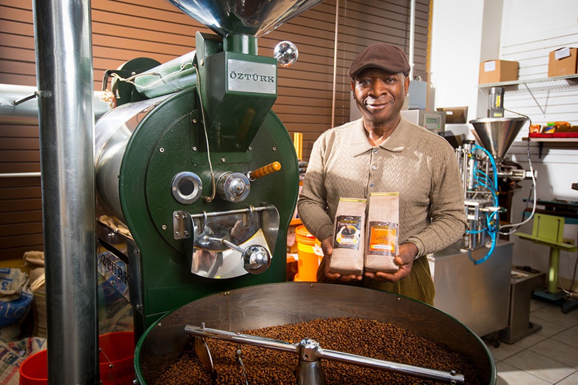 Larry Stitts, a coffee roaster who operates Golden Cup Coffee Co. and supplies coffee to UB cafes, stands in front of roasting machine and holds bags of coffee. 