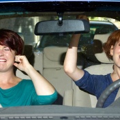 Two women in a car, one is plugging her ears while the other belts out a tune. 