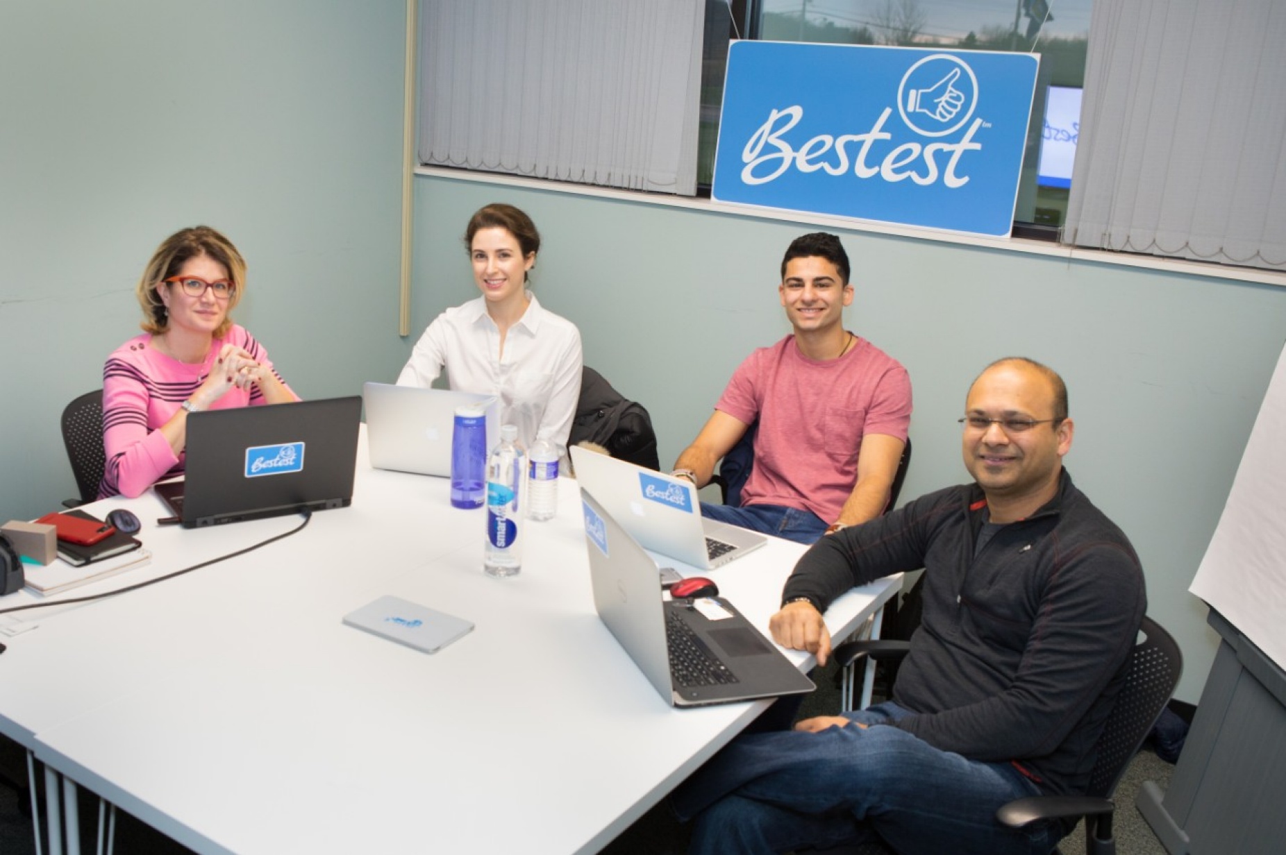 Bestest app leadership group seated around a table with Bestest logo behind them: Kelly Helmuth, chief growth officer; Rachel Shatkin, product development; Vick Goel, co-founder; and Lalit Goel, co-founder and CEO.