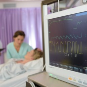 Closeup of a hospital alarm; in the background are the patient in bed, family member by her side and doctor standing with clipboard. 