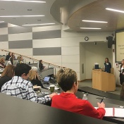 Ologie staff members Erin Burk and Carolyn Kent stand at a podium as they make a PowerPoint presentation (slide on giant screen behind them) in a lecture hall in Kapoor Hall. 