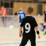 A young boy stands in his uniform watching an indoor soccer game. 