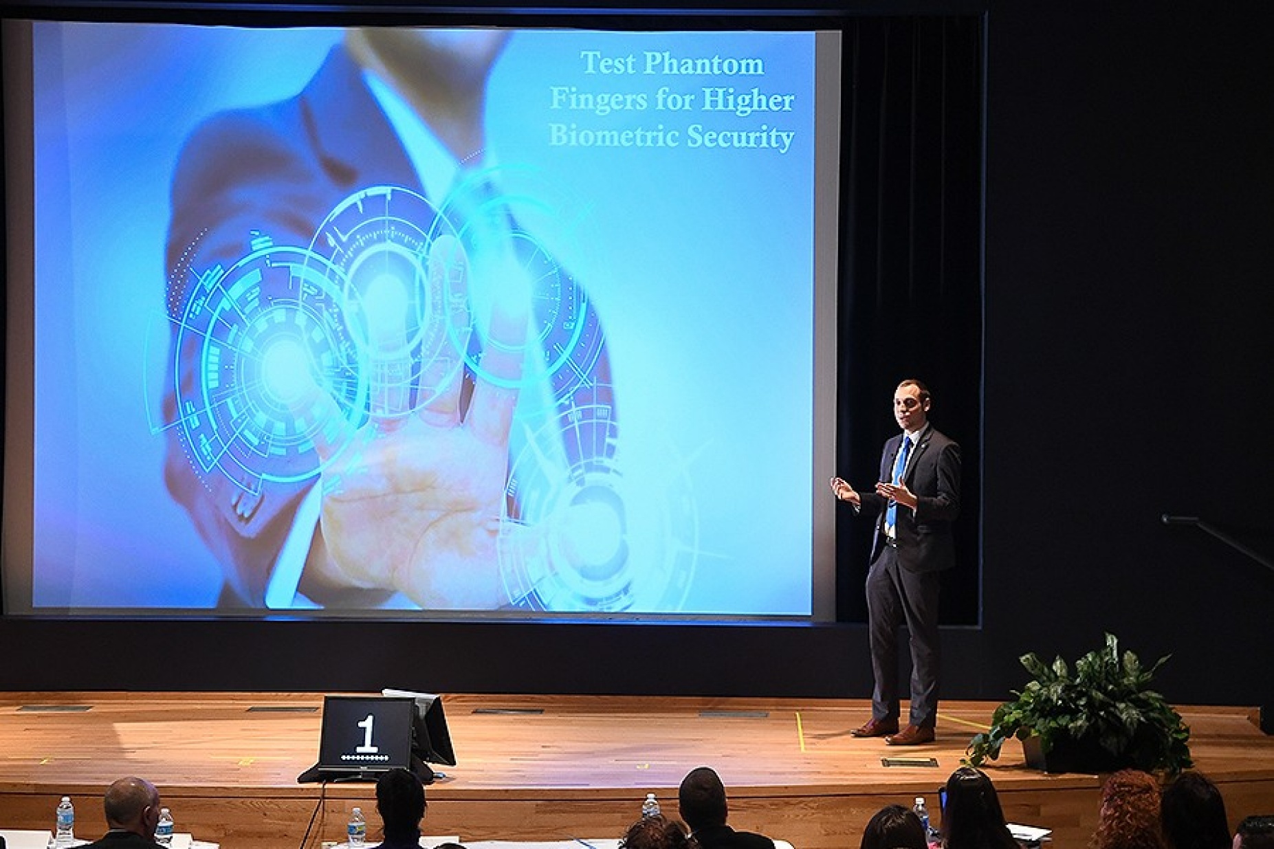 Phil Schneider makes his winning three-minute thesis pitch. He's standing in front of a giant screen that features the name of his pitch and his hand, with the ends of the fingers illuminated.