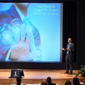 Phil Schneider makes his winning three-minute thesis pitch. He's standing in front of a giant screen that features the name of his pitch and his hand, with the ends of the fingers illuminated. 