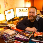 photo of Lance Rintamaki at his desk in his office, piles of books on the desk and the word SEX prominent on a computer screen behind him. 