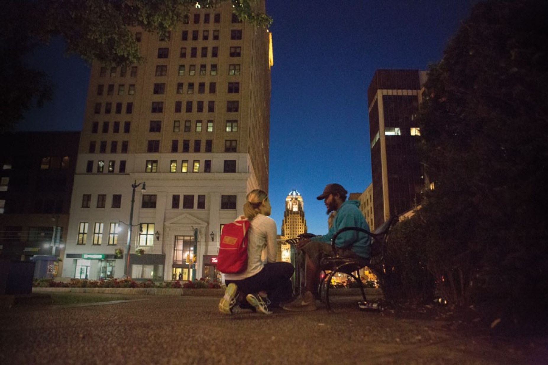 Kneeling medical student talks to a homeless person sitting on a bench in downtown Buffalo. city hall is in the background. 