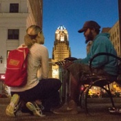 Kneeling medical student talks to a homeless person sitting on a bench in downtown Buffalo. city hall is in the background. 