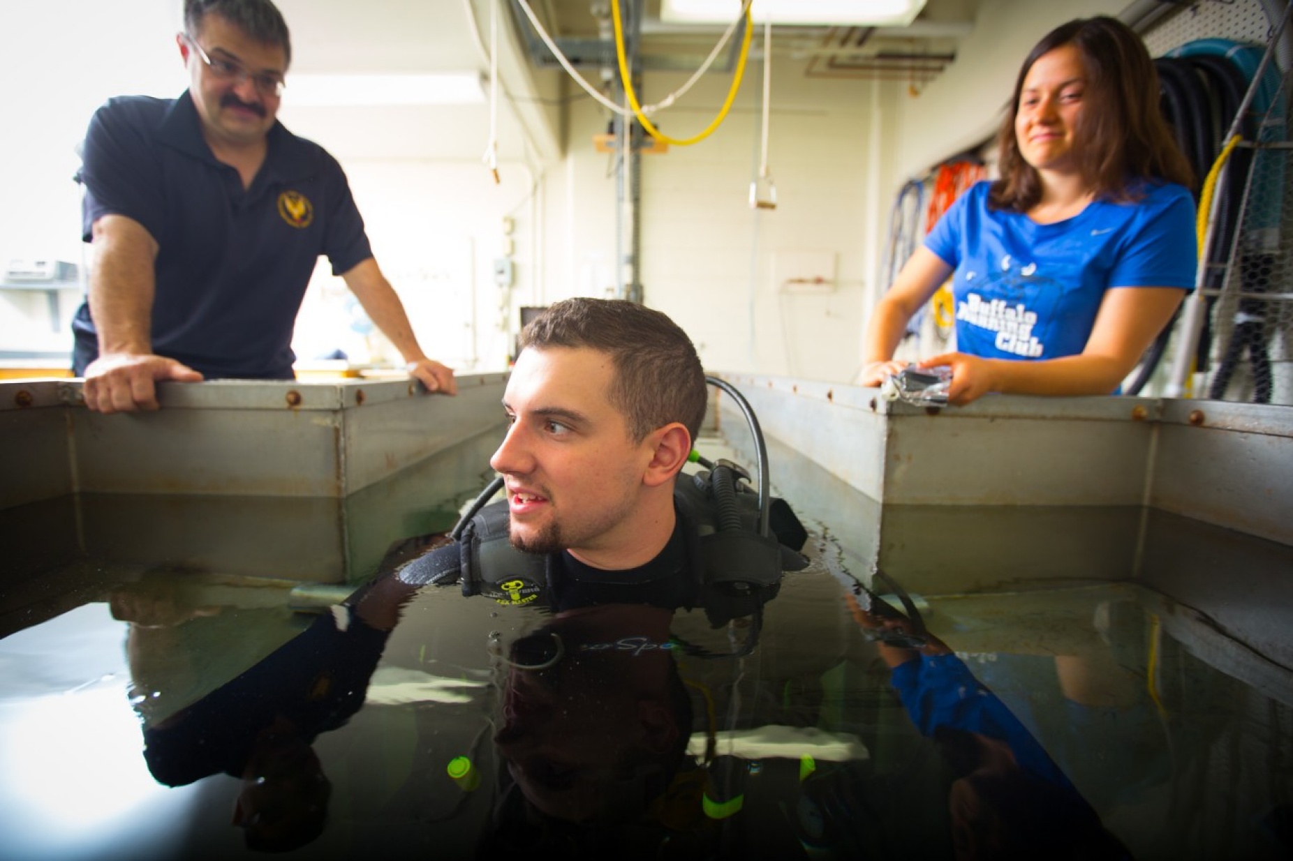 Dave Hostler looking on while a diver wearing a wetsuit crouches in a small pool. 