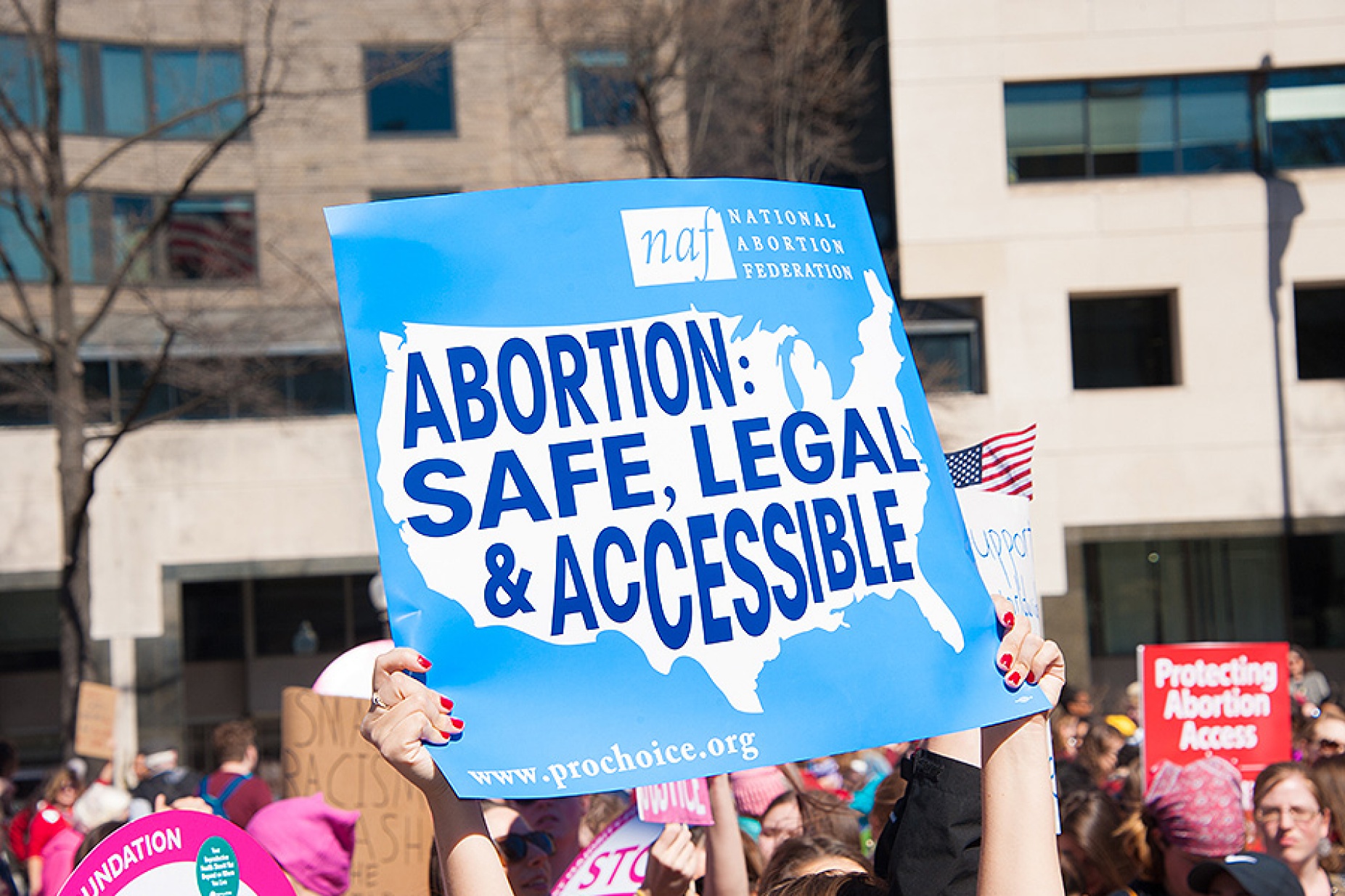 A woman's hands hold up a blue sign featuring a map of the U.s. and the words Abortion: Safe, legal & accessible. 