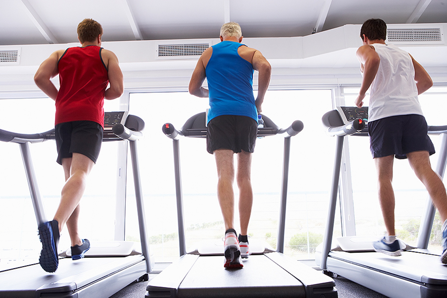 Back View Of Group Of Men Using Running Machines In Gym.