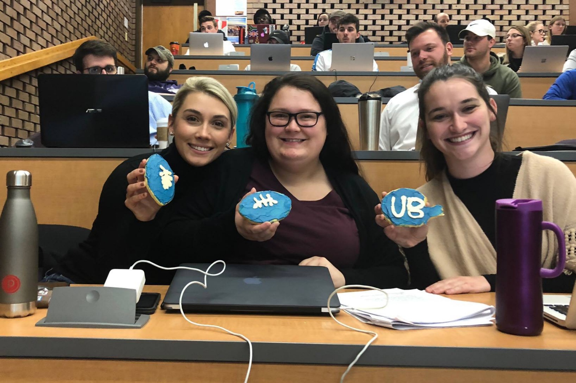 3 sports law students holding cookies shaped like footballs with UB Logo. 