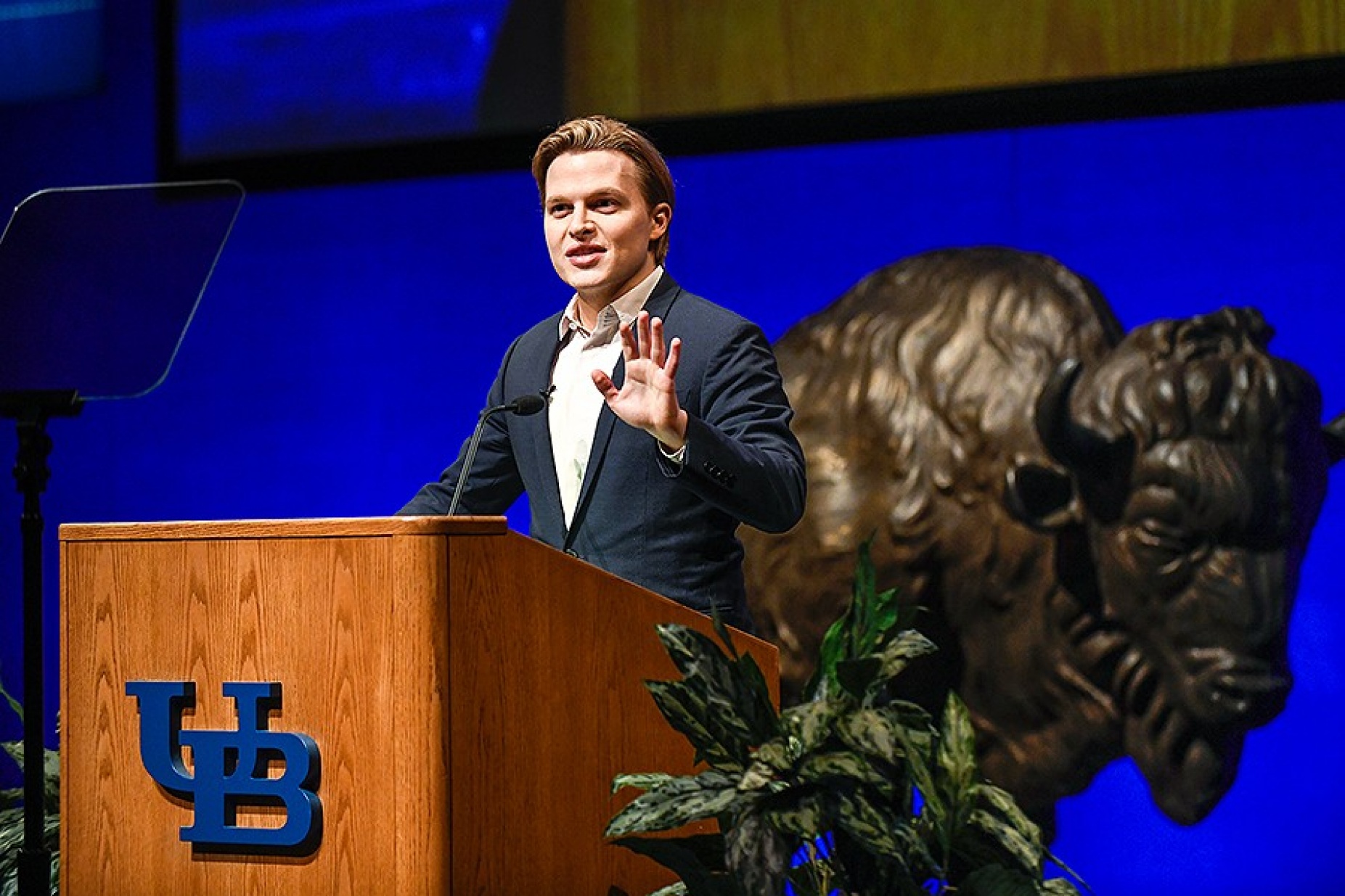 Ronan Farrow at the podium on the stage of the mainstage in the center for the arts, buffalo statue behind him. 