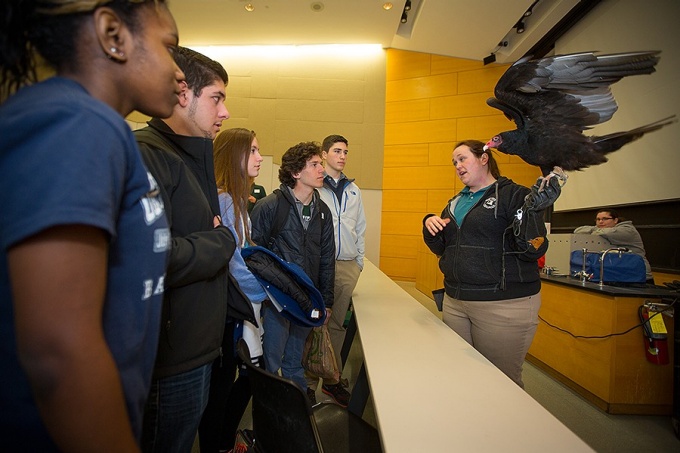 Students listen to a presentation while a turkey vulture perches on the presenter's arm. 