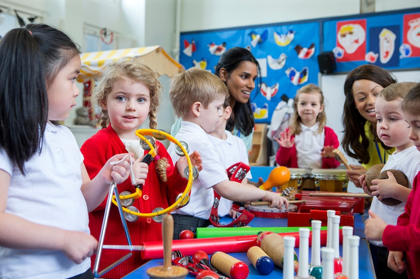 Young children in a preschool classroom, a girl plays a tambourine, a boy holds a ball. 