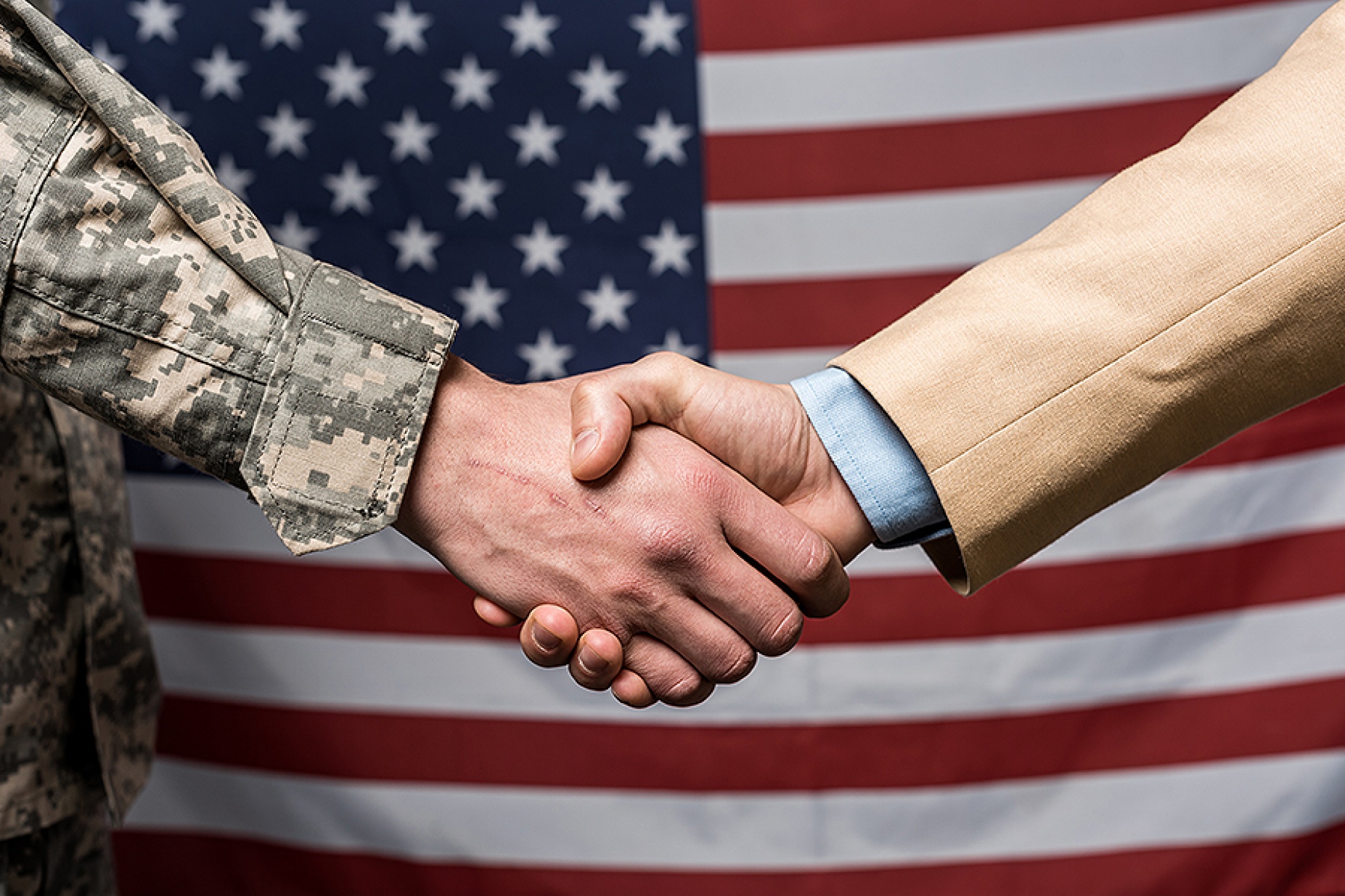 Man dressed in camo and a civilian in a suit shaking hands in front of an American flag. 