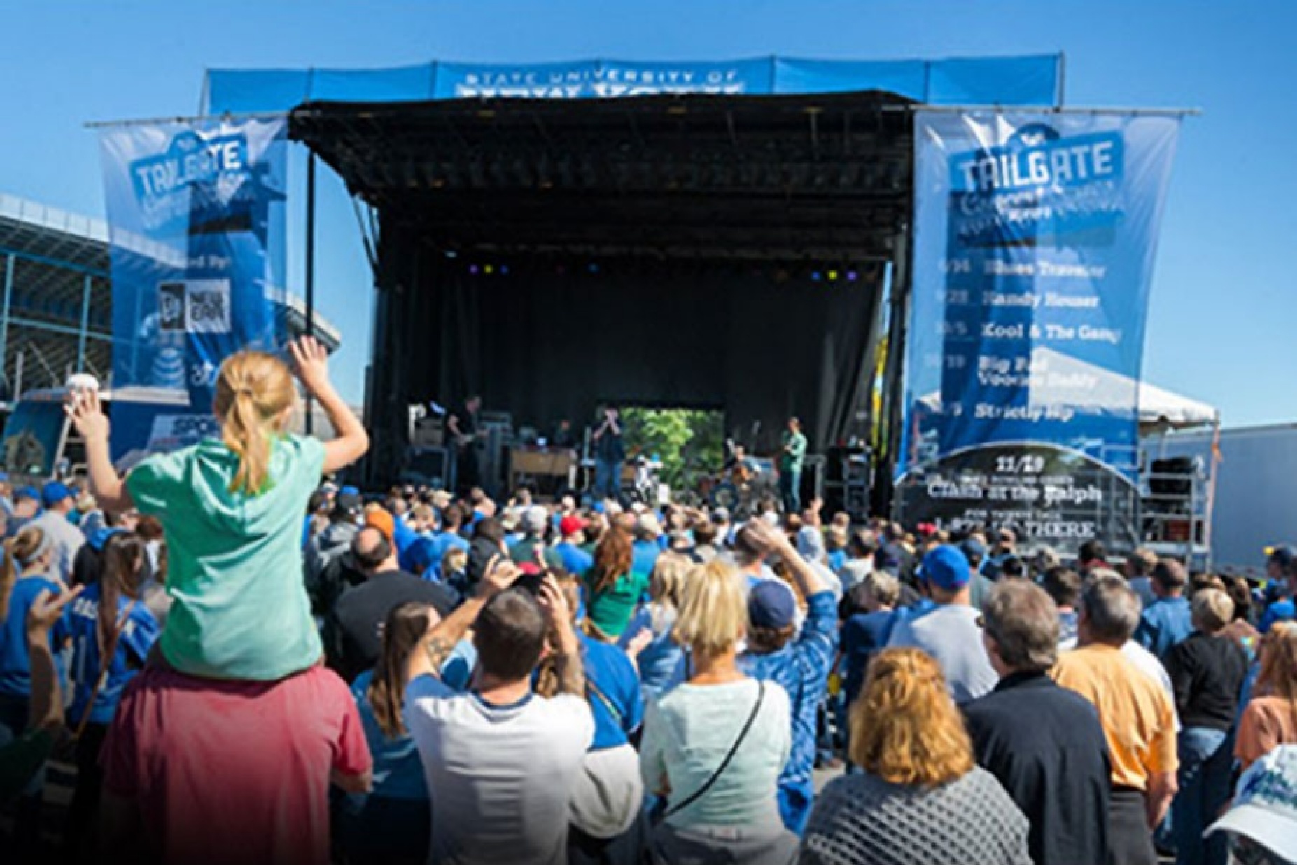 View looking from the audience's perspective into the stage and performers at a tailgate concert. A young girl sits on the shoulders of a man. 