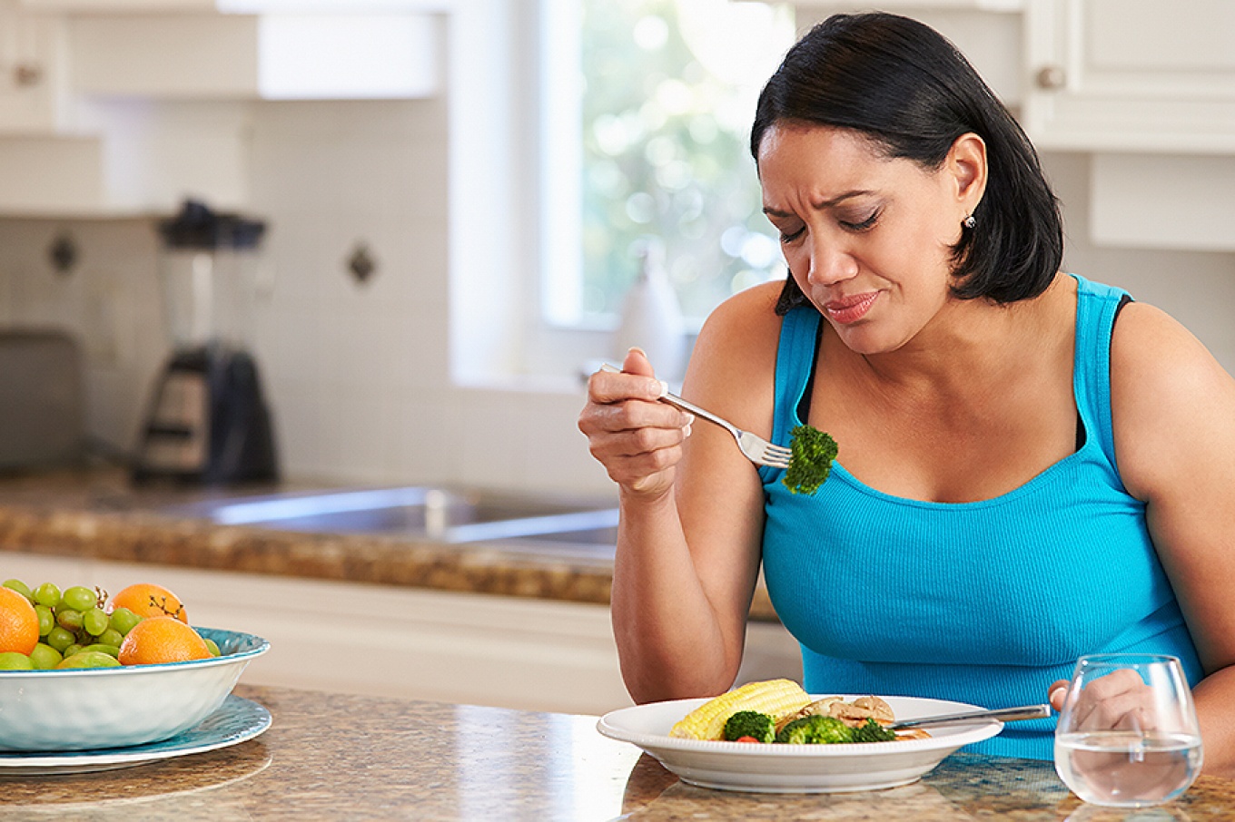 A frustrated- or disappointed-looking woman eating a healthy meal including broccoli in a kitchen setting. 