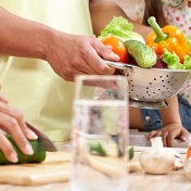 Meal preparation featuring a colandar full of vegetables and a cucumber being cut. 