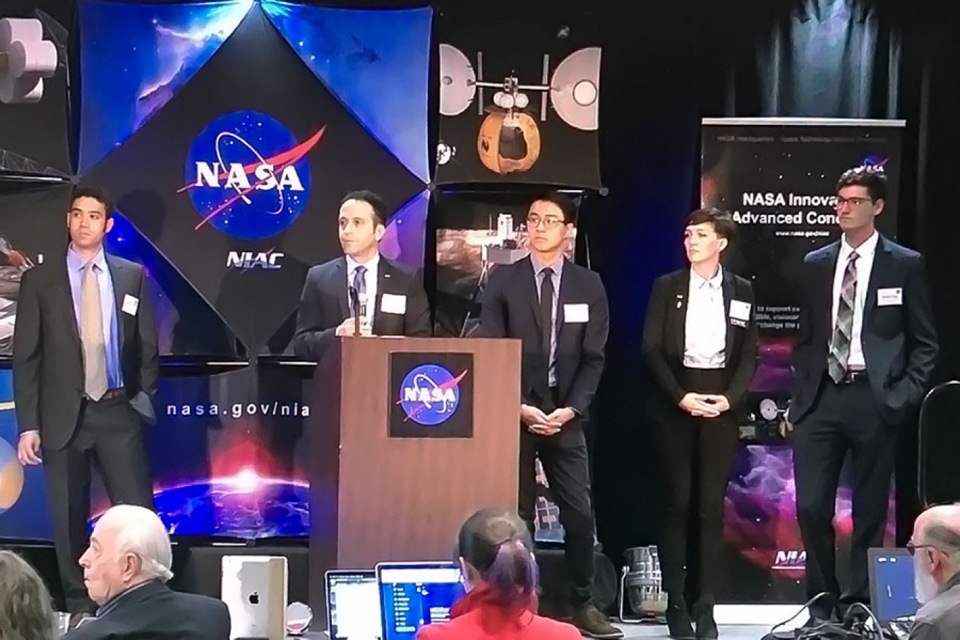 Javid Bayandor stands at podium at NASA event. At his side are student researchers. 