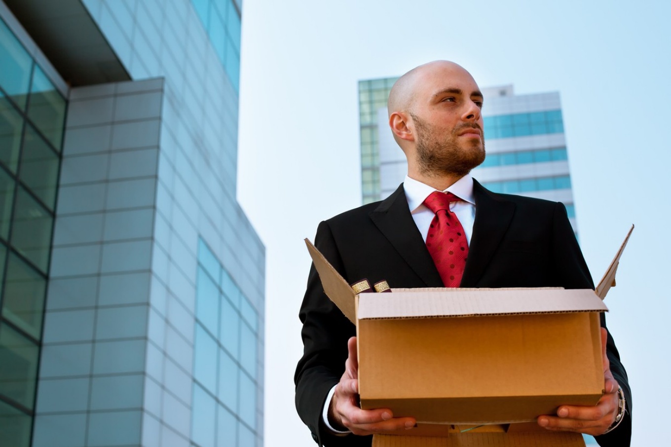 A man in a suit holding a box of office supplies, office building in the background. 