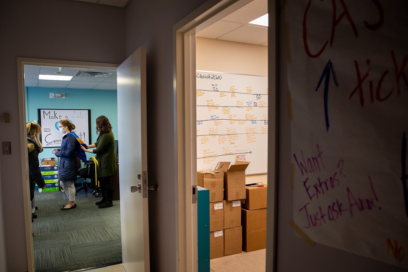 Zoom image: Anne Mecca (center) demonstrates how to position a graduation hood on student Cassondra Lyman, while Kayla Horisk looks on. In her office, at right, remnants of the 2020 year remain, with boxes of the outdated programs and a timeline still on the whiteboard. 