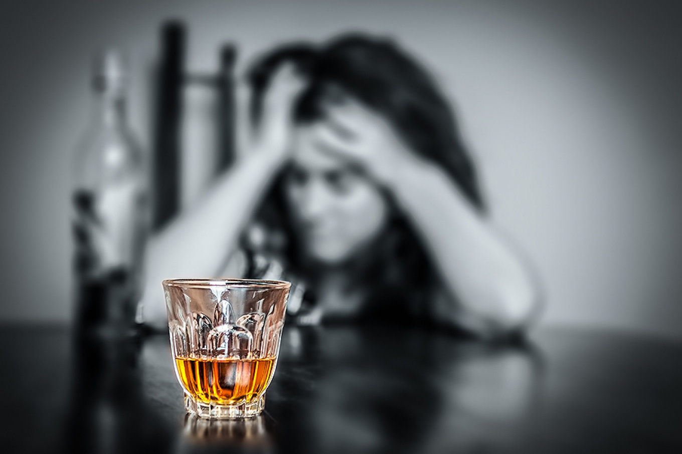 A glass of liquor in the foreground; woman with head in hands in background with bottle of liquor. 