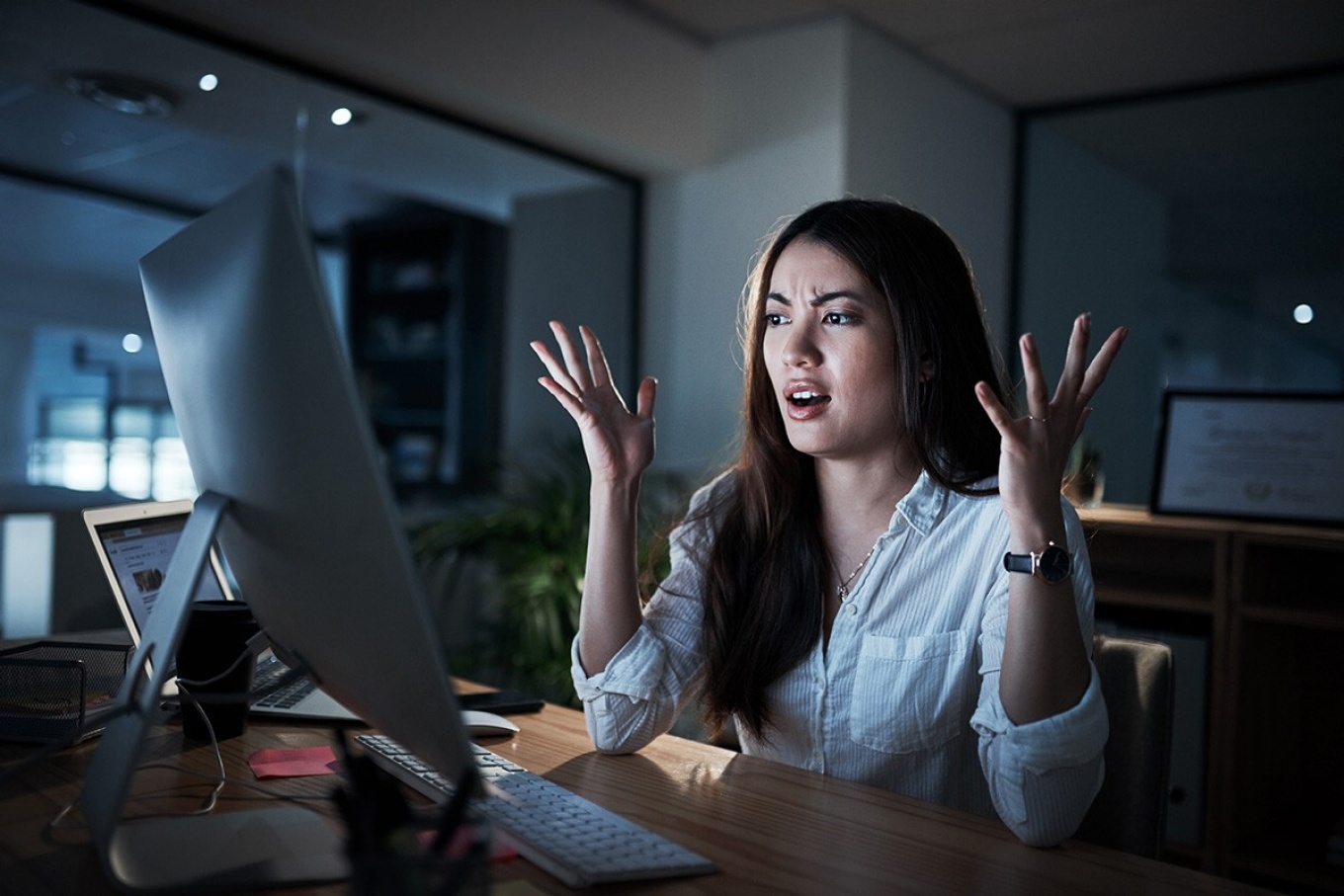 Young woman sitting at computer screen with hands in the air in frustration. 